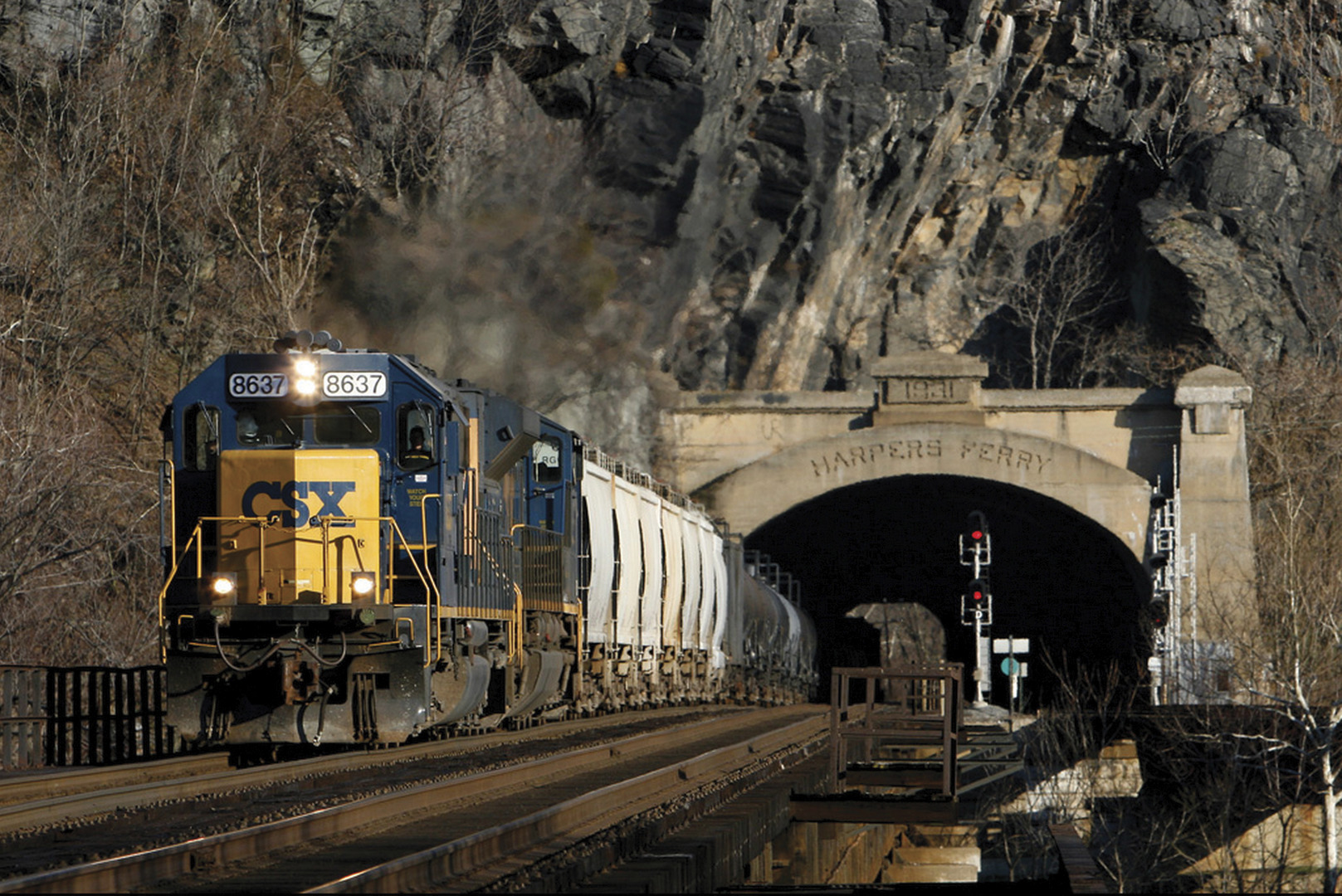A yellow and blue CSX train on a track emerges from a tunnel that cuts through a mountain