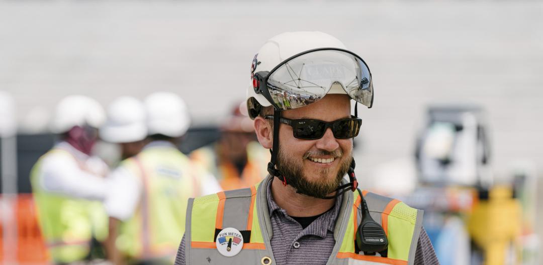 A Clark team member wearing personal protective equipment (PPE) - including a yellow safety vest, sunglasses, and a safety helmet - smiles at a jobsite in California