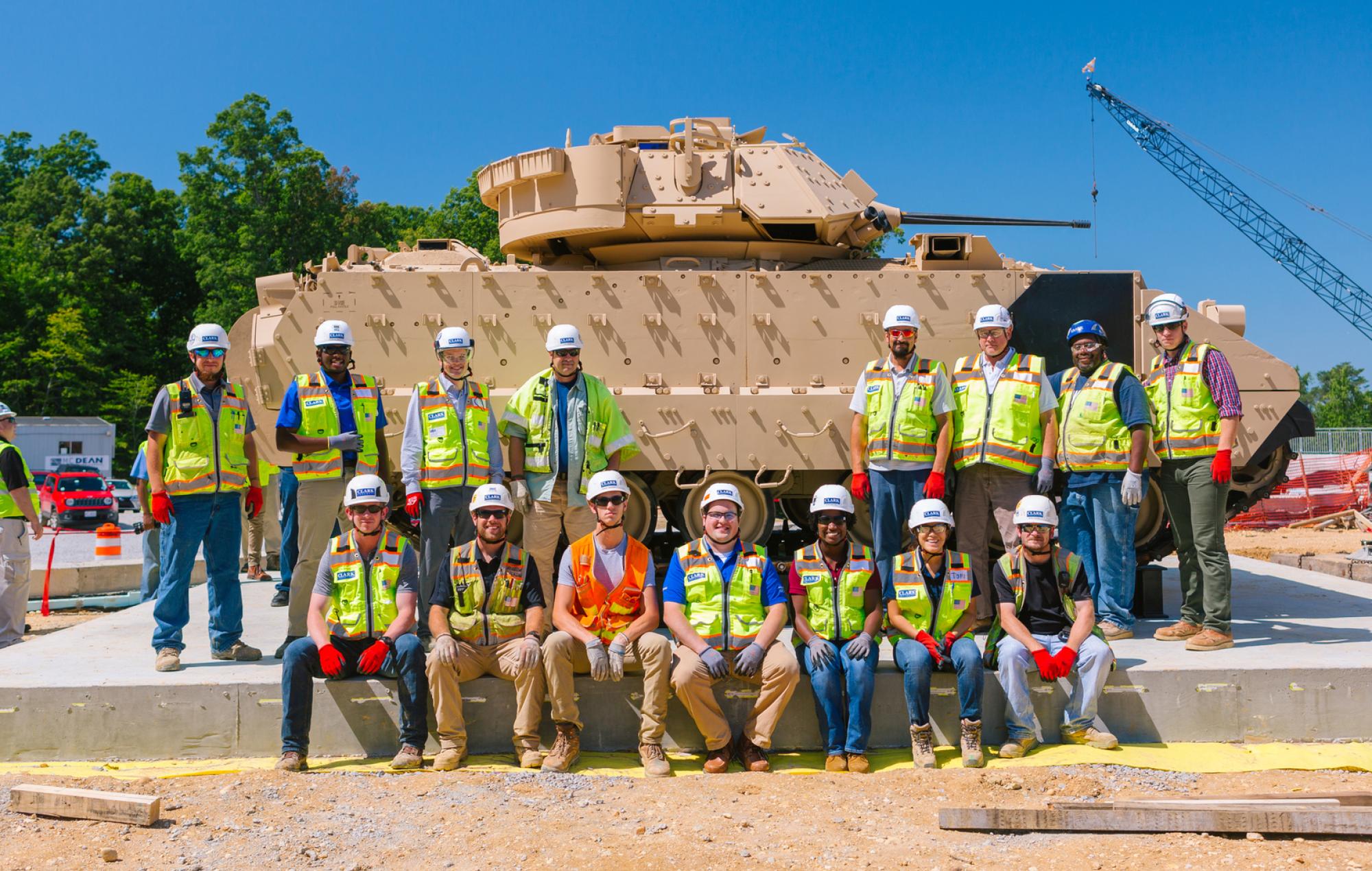 Clark employees in PPE pose in front of a sand-colored armored tank
